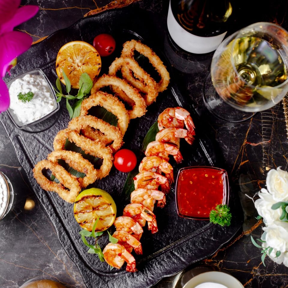 Fried calamari, grilled shrimp, and sauces on a black platter with wine.