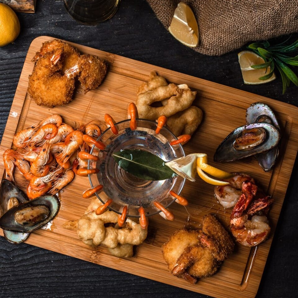 A wooden board with fried shrimp, mussels, and a glass of water with shrimp on the rim.
