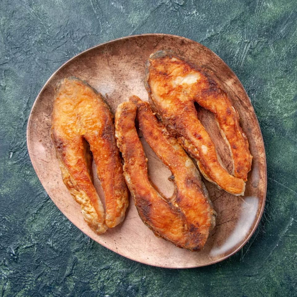 Three golden-brown fried fish fillets on a rustic copper plate, against a dark green textured background.